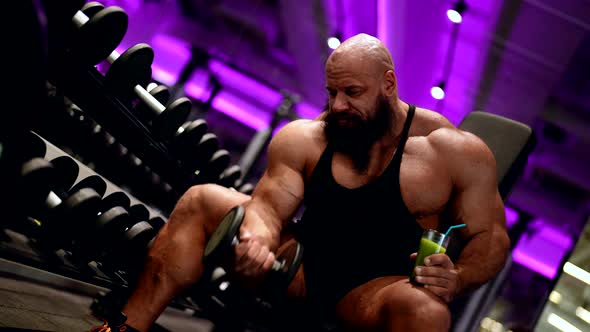 Powerlifter is Training Muscles Lifting Dumbbell and Drinking Juice with Protein in Gym alt