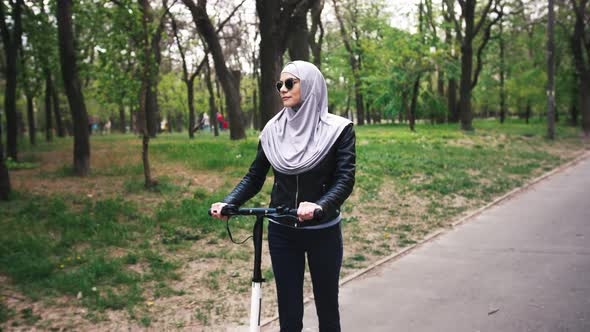 Portrait of Modern Muslim Woman in Sunglasses with a Hijab Riding Escooter in the Park Slow Motion