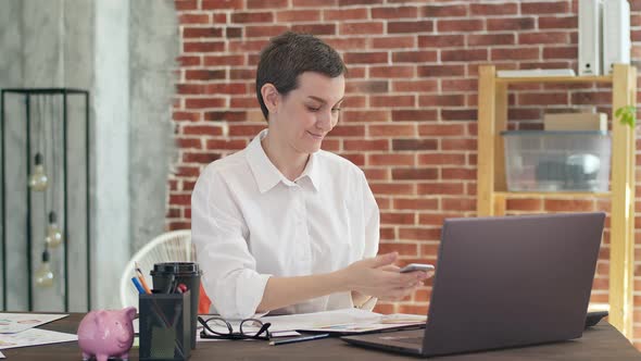 Nice Smiling Woman with a Short Haircut and a White Shirt Uses Phone and Prints on Laptop While alt