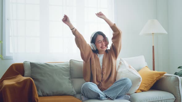 Happy asian woman wear wireless headphones having fun listen music in smartphone and sit on sofa alt