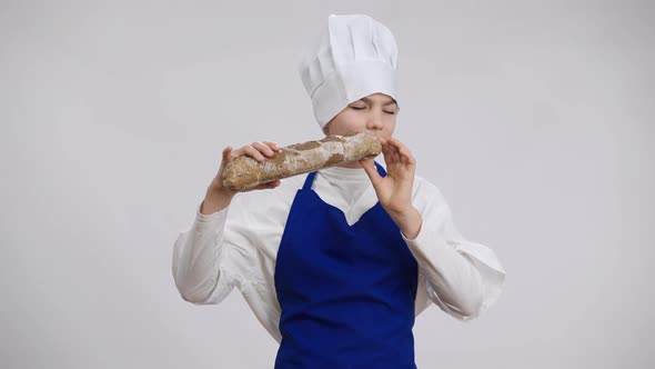 Happy Caucasian Boy in Chef Uniform Enjoying Smell of Freshlybaked Baguette Biting Tasty Food alt