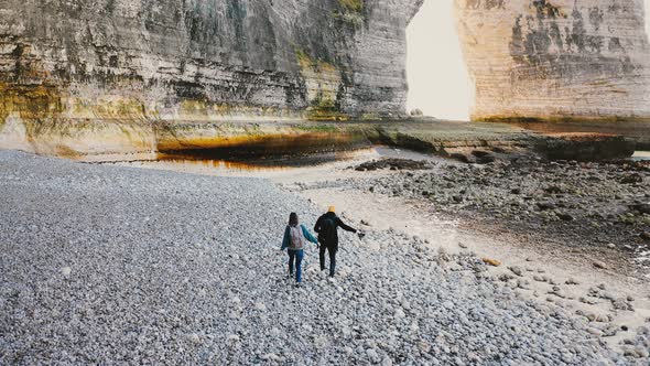 Drone Camera Follows Happy Tourist Man and Woman Walking Along Beautiful Big Pebble Beach To alt