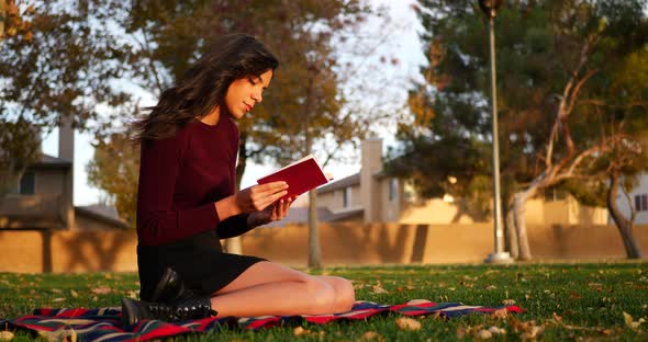 A gorgeous young hispanic woman student reading a book or novel outdoors on a college university cam alt