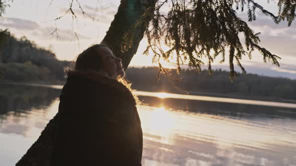 Woman Sits on the Shore of a Lake Near the Water in the Rays of the Setting Sun