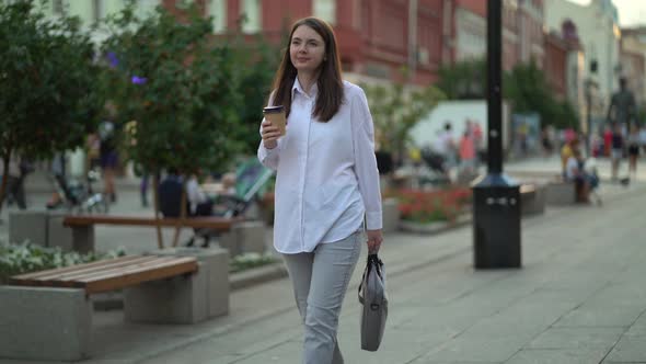 Modern Businesswoman with a Cup of Coffee and a Laptop in Her Hands Walks Along the Sidewalk on the