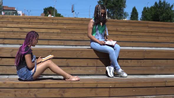 Two Girls are Reading a Book Sitting on a Wooden Bench in the Park in Sunny Weather alt