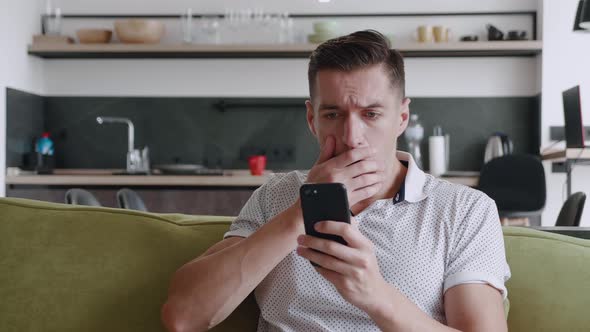 Shocked Man Reacting Message on Smartphone Covers His Mouth in Fright Sitting on Sofa at Home Living alt