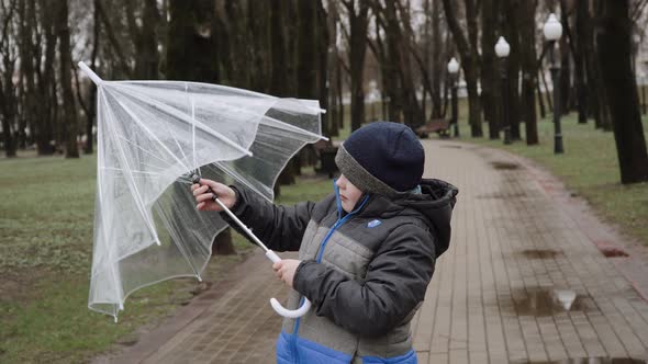 Caucasian Boy in Cold Rainy Weather Looks Up and Folds Transparent Umbrella alt