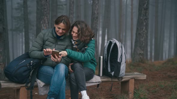 Two Young Women Using a Smartphone While Sitting on a Bench in a Forest  alt
