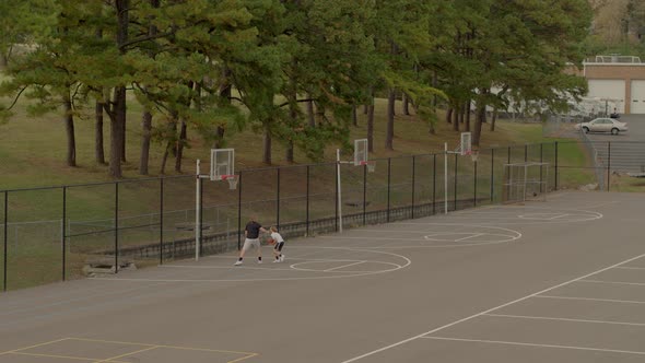 two teen boys play one on one basketball on an outdoor court alt
