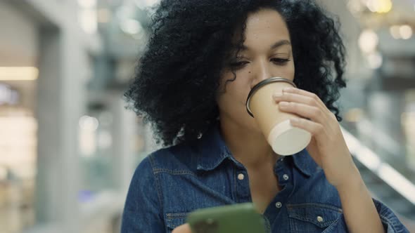 Happy Fashion African American Woman Texting on Her Phone and Drinking Coffee While Standing in Mall alt