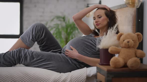 Young Charming Pregnant Woman in Pajamas Lying on Bed Stroking Belly Thinking alt