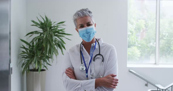 Portrait of caucasian female senior doctor wearing face mask standing with arms crossed in hospital alt