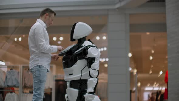 A Man Stands with a Robot Bot and Asks Him Questions and Asks for Help By Clicking on the Screen on alt