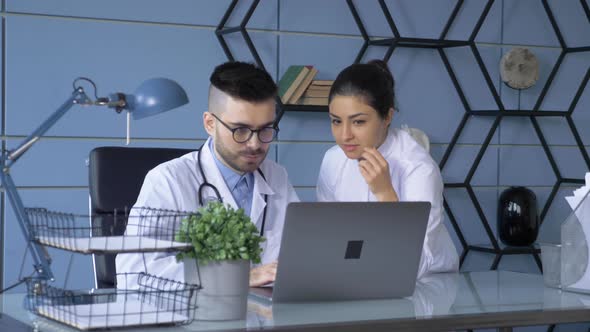 Male doctor using laptop and talking to female colleague at medical clinic. Young man looking alt