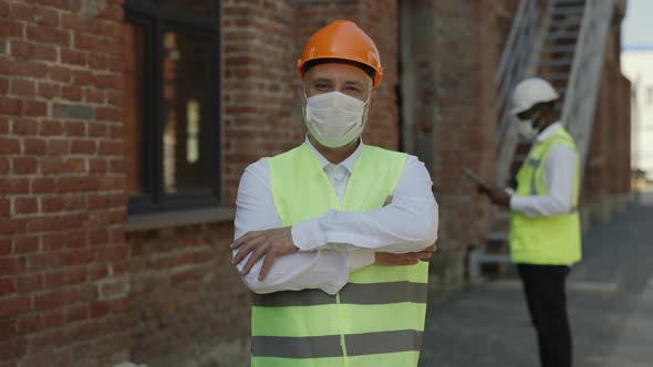 Builder in Mask Posing with Crossed Arms at Building Site alt