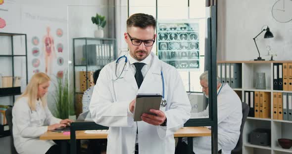 Adult Bearded Medic in white Coat which Using Tablet pc in Clinic Office alt