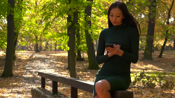A Young Asian Woman Sits on A Wooden Railing in A Park and Works on A Smartphone alt