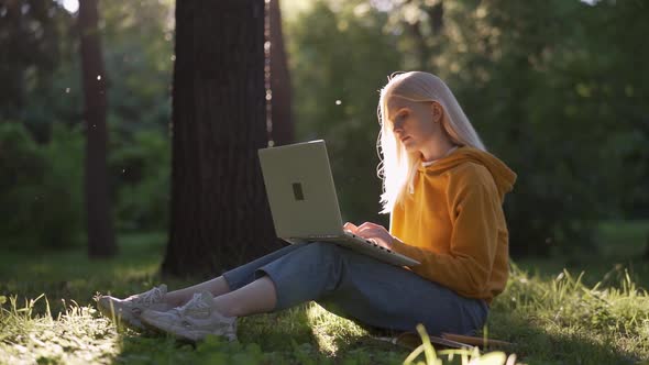 A Tender Young Woman with Blond Curls Sits on the Green Grass with a Laptop
