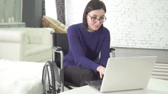 Young Disabled Woman with Glasses in a Wheelchair Chair with a Laptop at Homesmiling alt