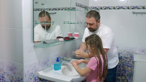 Dad teaches a little girl how to wash their hands with soap over a sink with running water
