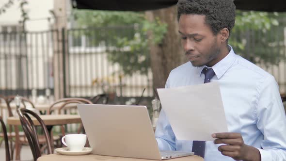 African Businessman Working on Contract and Laptop in Outdoor Cafe alt
