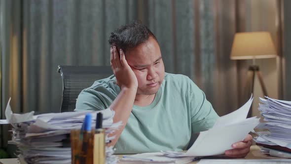 Close Up Of Fat Asian Man Yawning And Putting Hand On Cheek While Working Hard With Documents alt