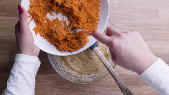 Hands Adding Grated Carrots with Tablespoon into Carrot Cake Dough. - Top Down Shot alt