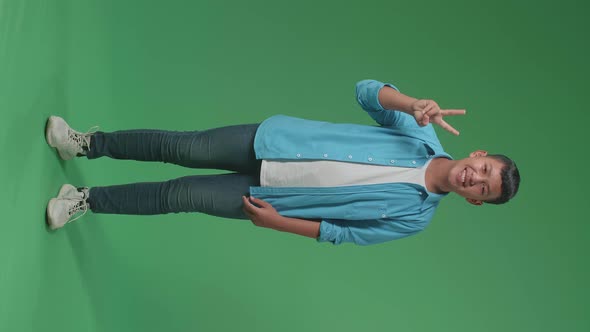 Full Body Of Smiling Asian Boy Showing Gesture Peace While Standing On Green Screen In The Studio alt