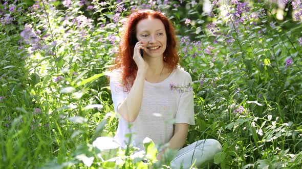 Young Red-Haired Woman Talking on a Mobile Phone Among Flowers 