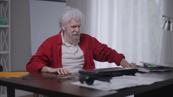 Senior Bearded Caucasian Disabled Man Closing Laptop Rolling Wheelchair to Window in Home Office alt