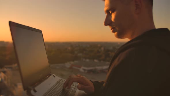 A Male Stockbroker Freelancer Stands on a Rooftop at Sunset with a Laptop and Types on a Keyboard alt