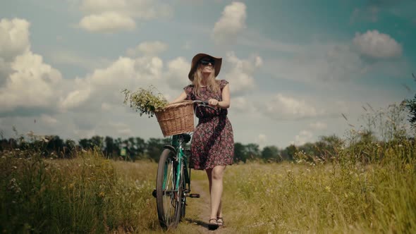 Tourist Girl Relaxing On Countryside Wildflower Field. Woman Cyclist Walking With Bike On Holiday. alt