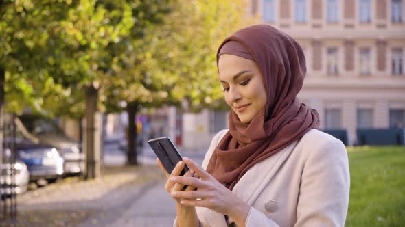 A Young Beautiful Muslim Woman Takes Selfies with a Smartphone in a Street in an Urban Area alt