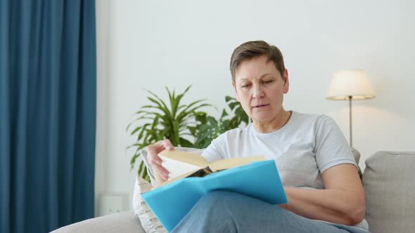 Relaxed Senior Woman Sitting on Sofa and Reading Book Indoors at Home alt
