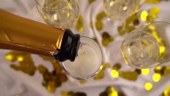 Pouring from a bottle of sparkling wine into glasses on a background of confetti. Slow motion. alt