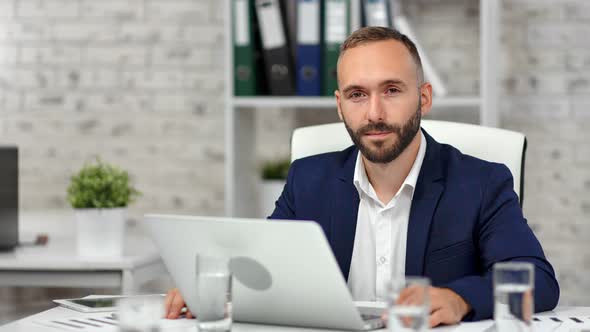 Portrait of Smiling Bearded Businessman in Suit Posing at Modern Light Office in Front of Laptop Pc alt