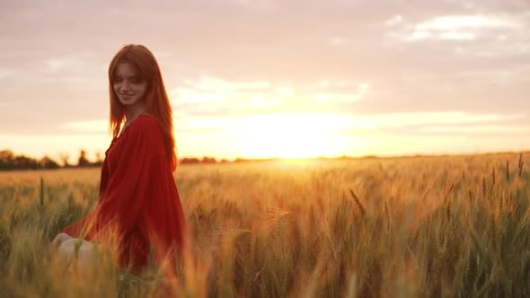 Romantic Redhead Girl in Dress Playing with Wheat Ears on Field at Sunset