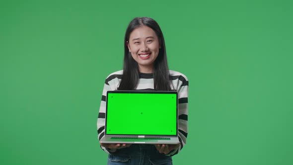 A Smiling Asian Woman Holding Green Screen Computer While Standing In Front Of Green Screen alt