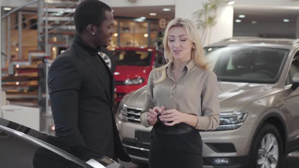 Pretty Caucasian Woman Giving Car Keys To the African American Man in Automobile Showroom alt