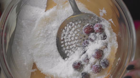 Slow motion overhead metal strainer sifting bright red cranberries and sugar in clear glass bowl on alt