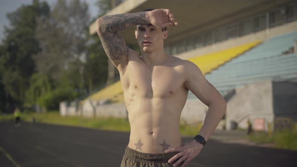 Middle Shot of Perspiring Sportsman Wiping Forehead with Tattooed Hand and Looking at Camera alt