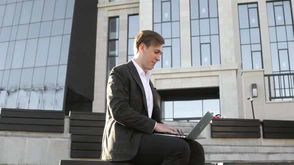 Businessman Startupper Typing on Laptop Sitting on Bench Near Office Building.