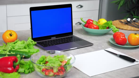 Blue Screen Laptop on Kitchen Table Near Vegetables and Cutting Board with Knife alt