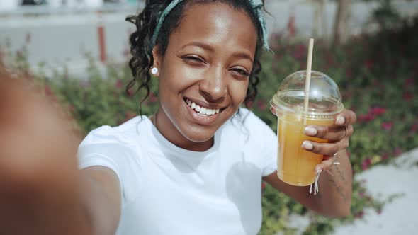 Smiling brunette African woman recording video and drinking juice alt