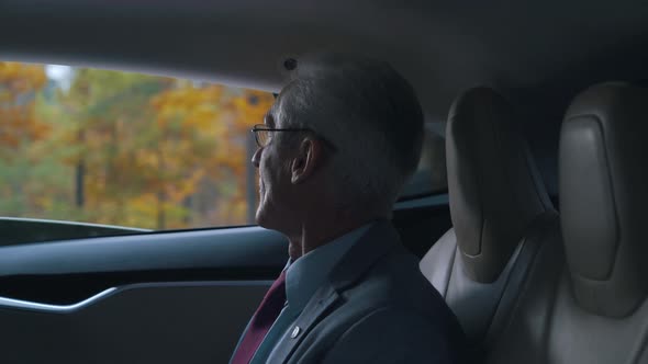 Old Businessman Rides with an Open Window Sitting in the Back Seat of a Car