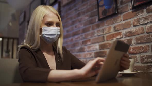 A Young Attractive Woman in a Medical Mask Sits in a Cafe and Scrolls the Tablet