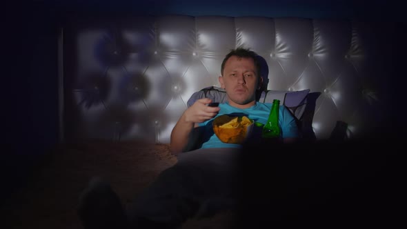 Man spends the evening lying in bed by the TV with a bottle of beer and a bowl of chips alt