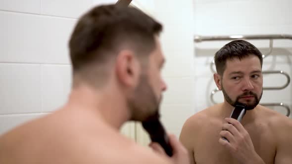 young caucasian man trimming his beard in bathroom, reflection in mirror