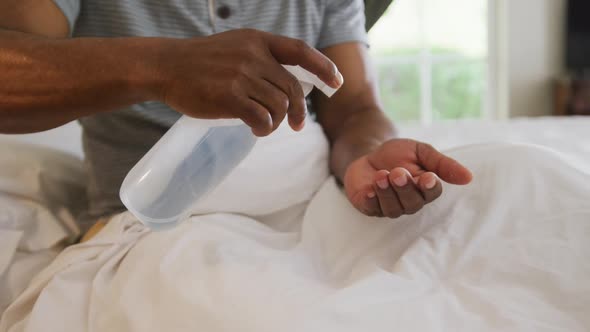 Mid section of african american senior man sanitizing his hands while sitting on the bed at home alt
